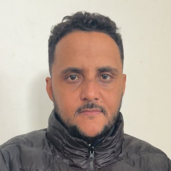 Islamic Center A man with short curly hair and a trimmed beard, wearing a black puffer jacket, stands against a plain off-white background. Islam