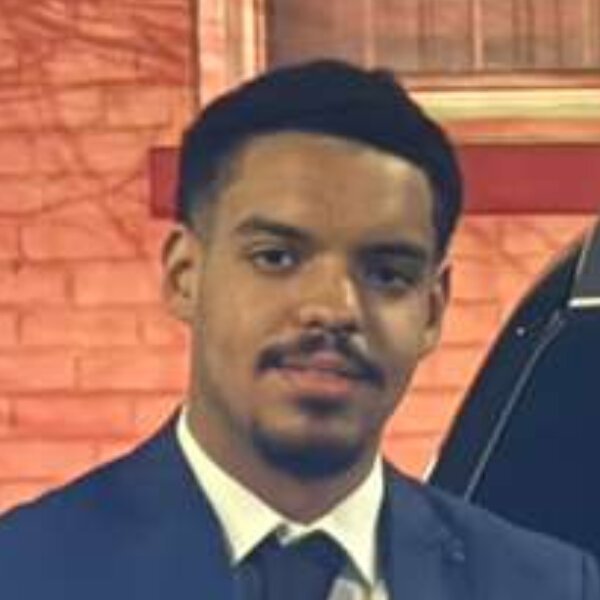 Islamic Center A man in a dark suit and tie stands in front of a brick wall and a parked vehicle, looking at the camera. Islam
